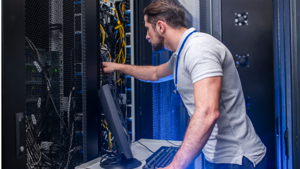 A man focused on his computer in a server room, surrounded by rows of servers and blinking lights.